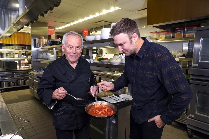 Documentary director David Gelb tastes food from a pot held by Wolfgang Puck in a kitchen.