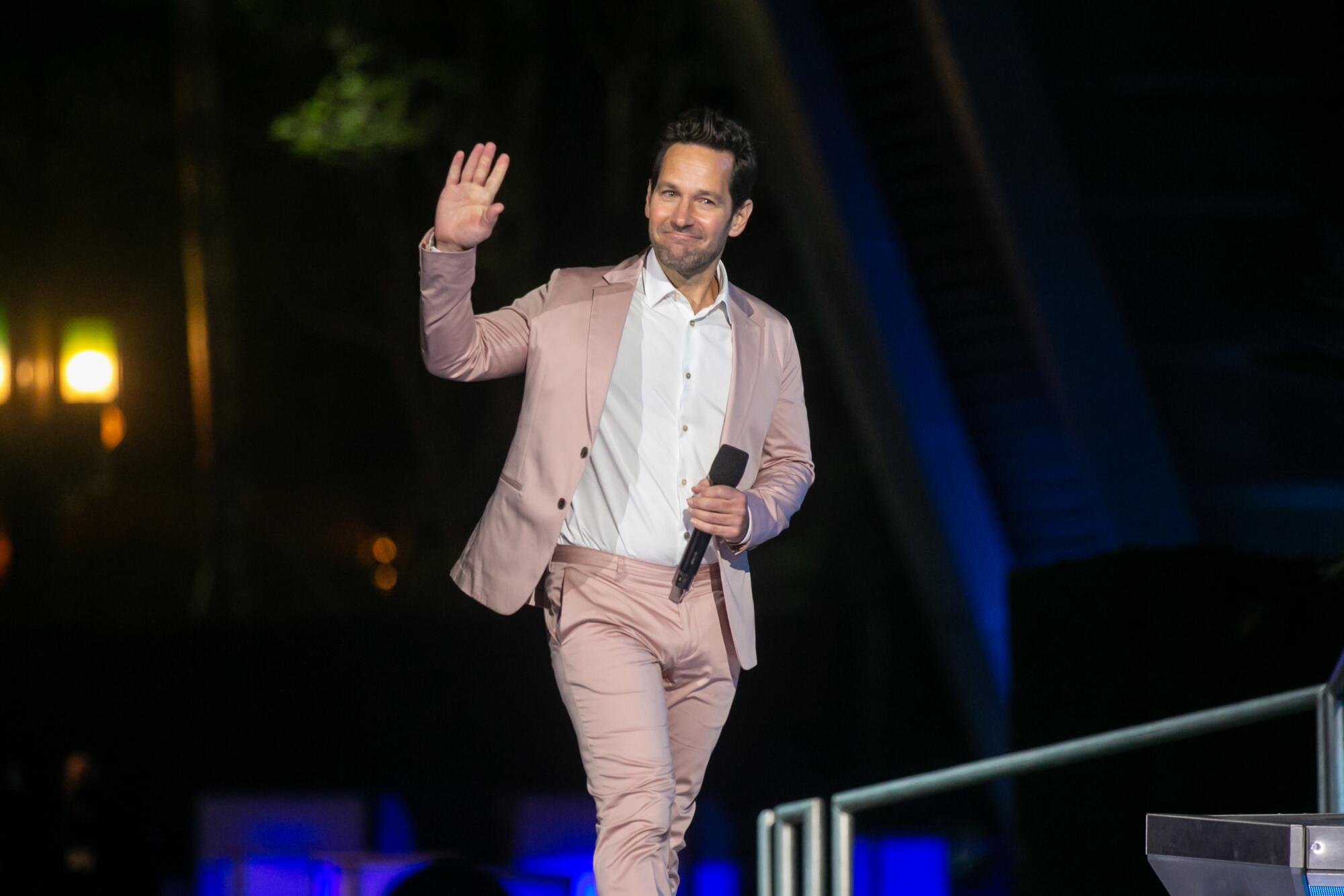 Paul Rudd, wearing a pink jacket and trousers, waves.