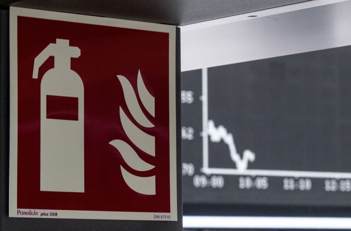 The display board with the curve of the DAX photographed behind a pictogram for a fire extinguisher in the trading room of the Stock Exchange in Frankfurt, Germany, Thursday, March 12, 2020. This was again triggered by concerns about the economic consequences of the coronavirus pandemic. For most people, the new coronavirus causes only mild or moderate symptoms, such as fever and cough. For some, especially older adults and people with existing health problems, it can cause more severe illness, including pneumonia. (Boris Roessler/dpa via AP)