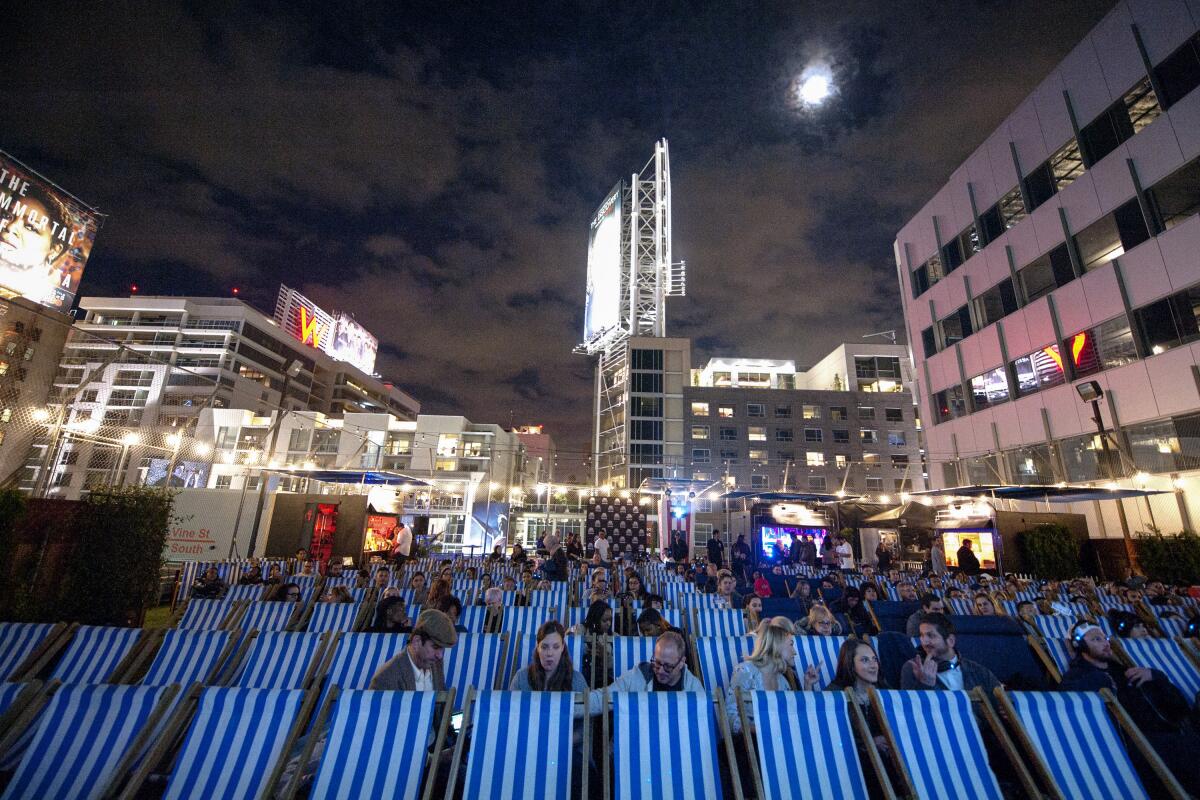 People sit in lounge chairs facing a movie screen, with tall buildings in the background.