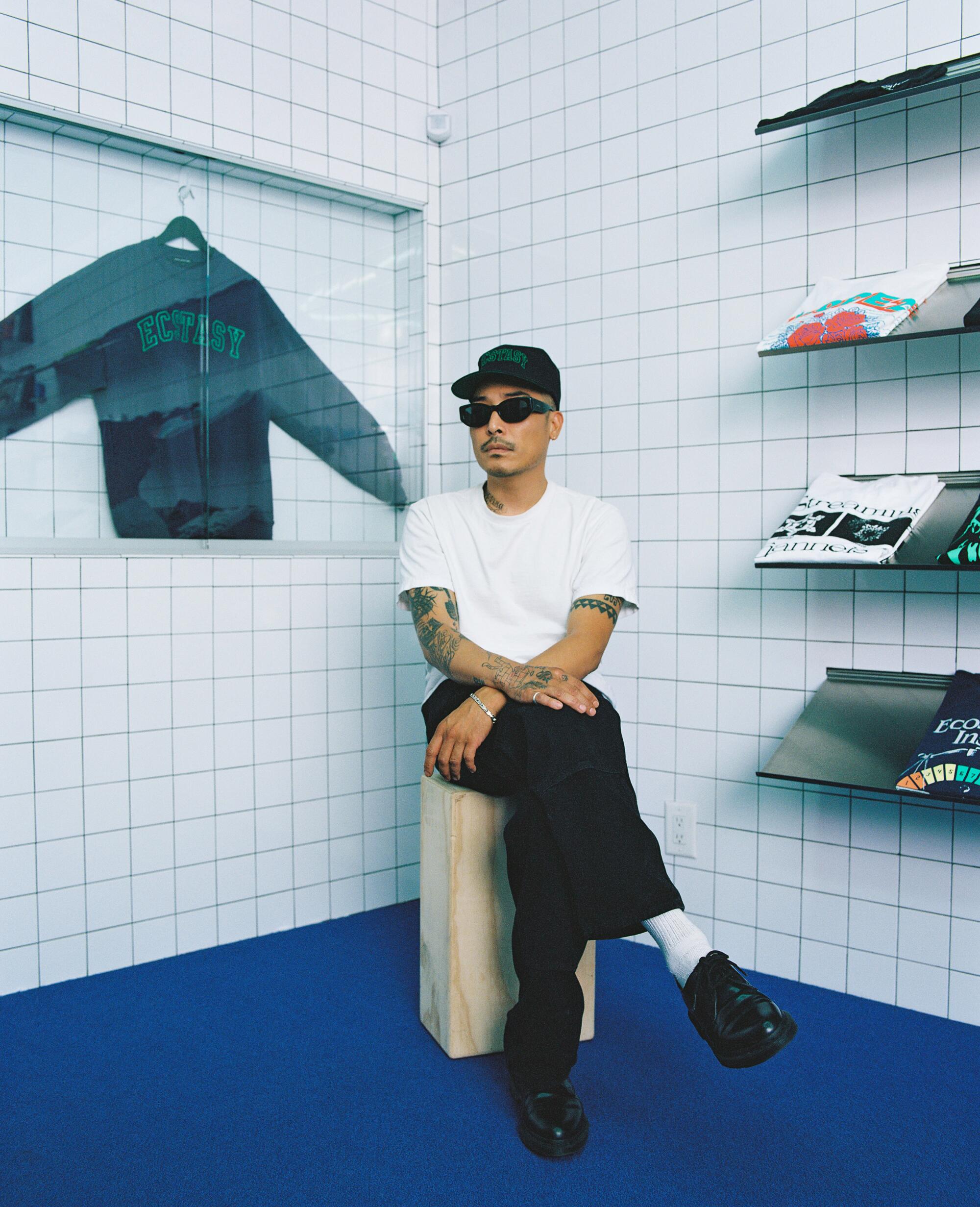 A man in sunglasses, white T-shirt and black pants sits in a store with white tile walls and a blue floor.
