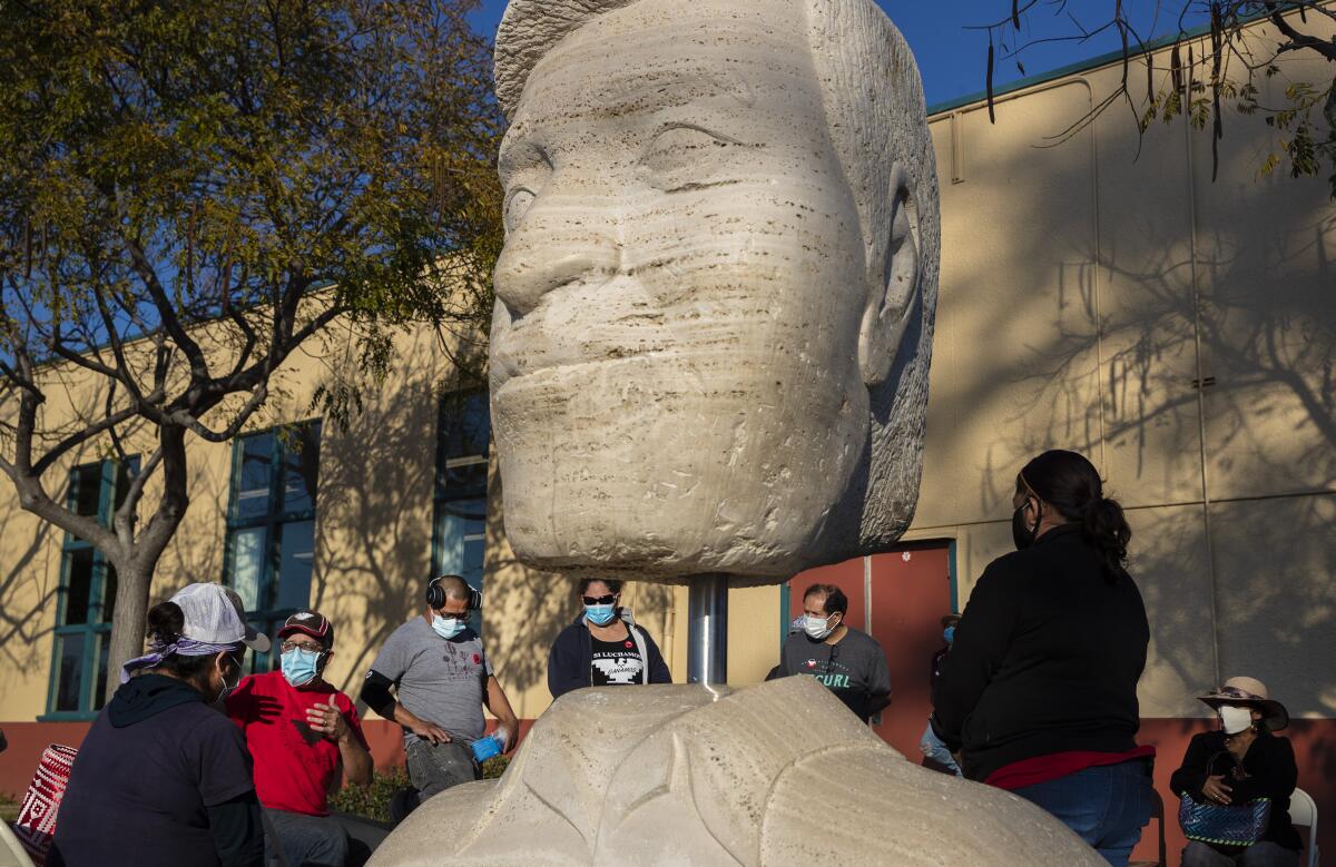 People in masks gather outdoors near a sculpture.