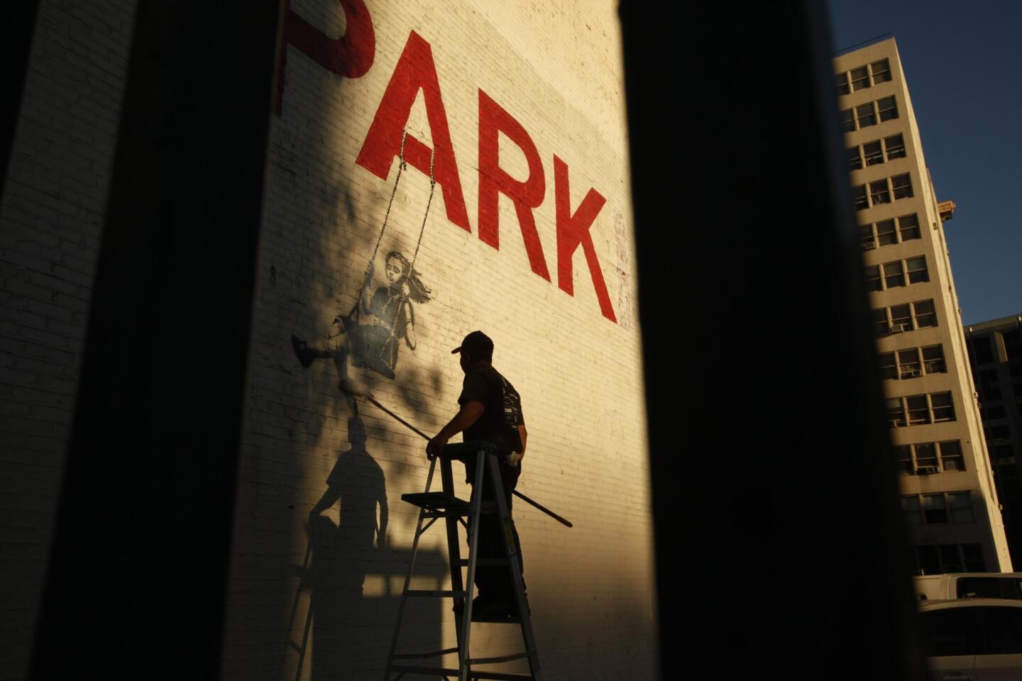 A young girl swings from the word "PARK" within Banksy's downtown Los Angeles mural. But an additional "ING" has been faded out by Banksy, turning the word "PARKING" into "PARK." Just down the block from this area, a downtown resident group is looking to find a little more than $6 million to transform a parking lot into a community park with a playground.