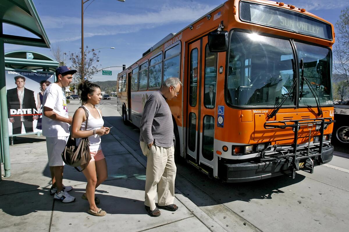 Three people standing on a sidewalk, waiting to board an orange bus