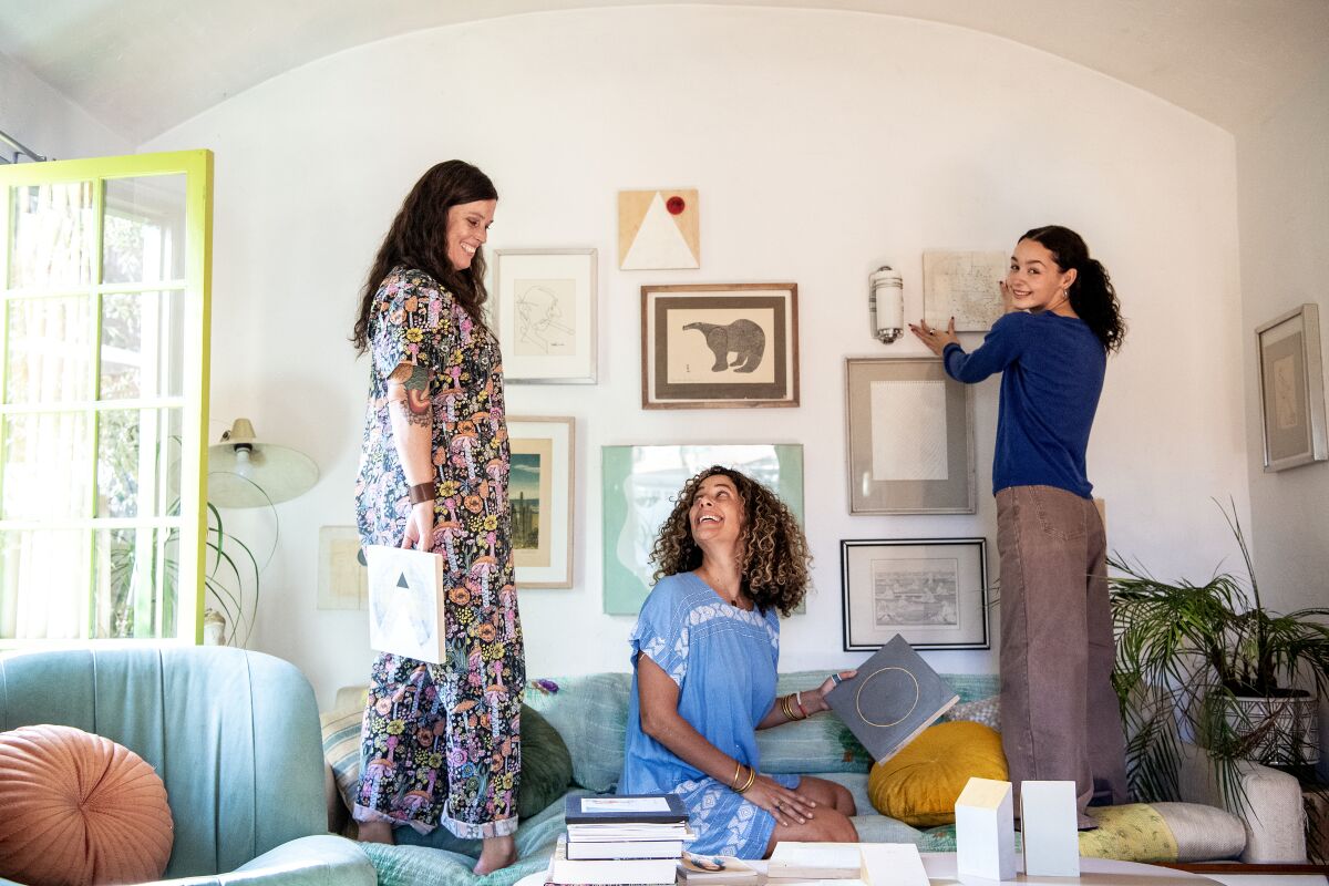 Two women and a teenage girl stand on a couch as they hang art in a living room.