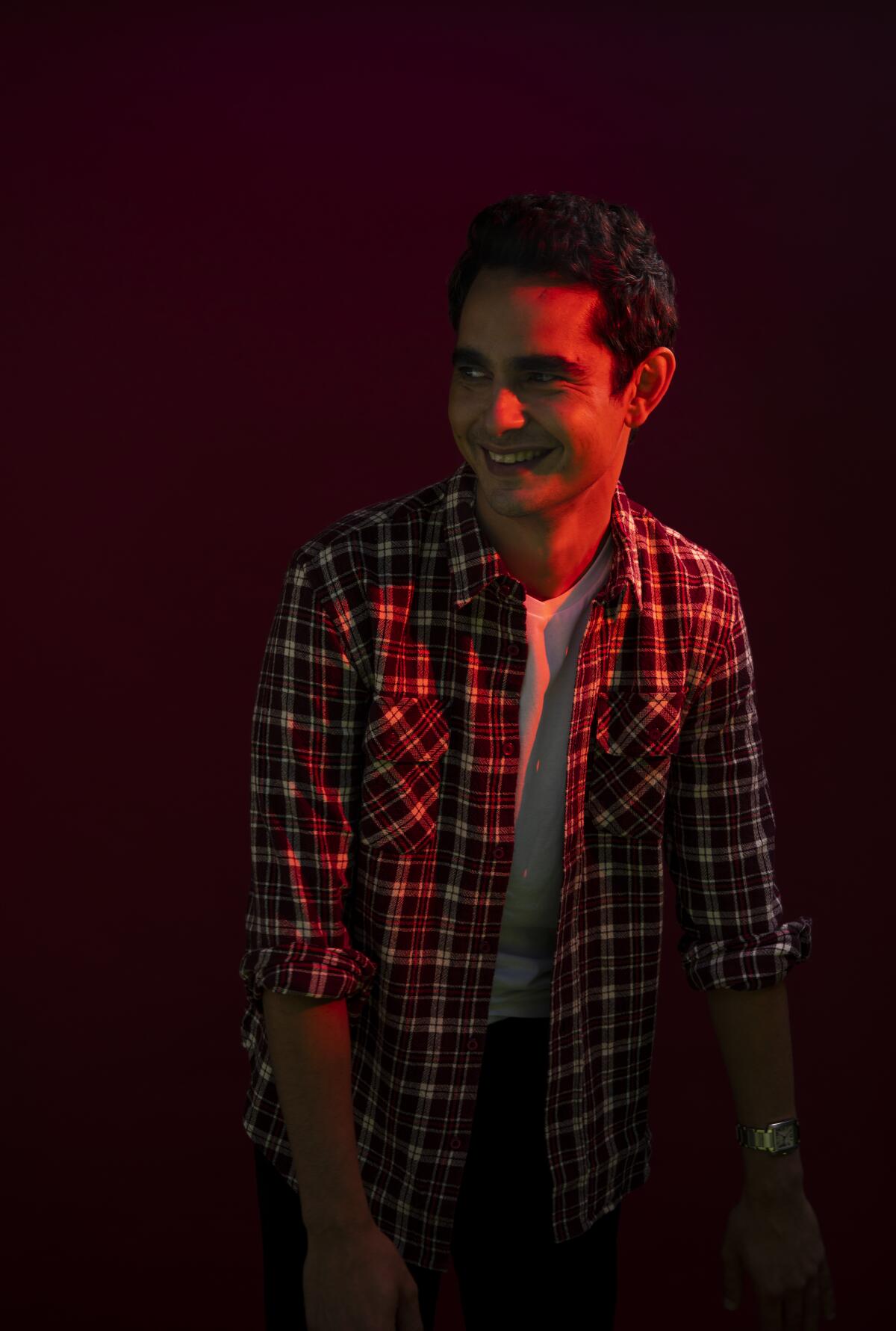 Max Minghella smiles while wearing a plaid shirt over a T-shirt.