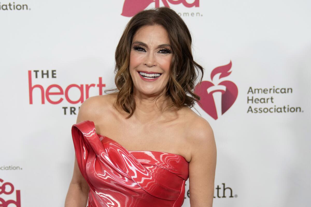 Teri Hatcher smiles in a shiny, strapless red dress with wavy shoulder-length brown hair against a white background