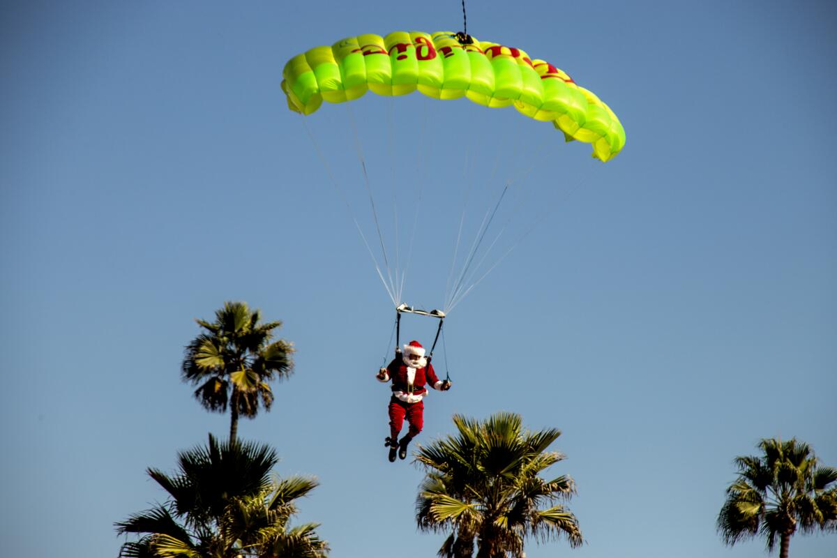 Above palm trees, a man in a red suit and white beard dangles from a parachute.