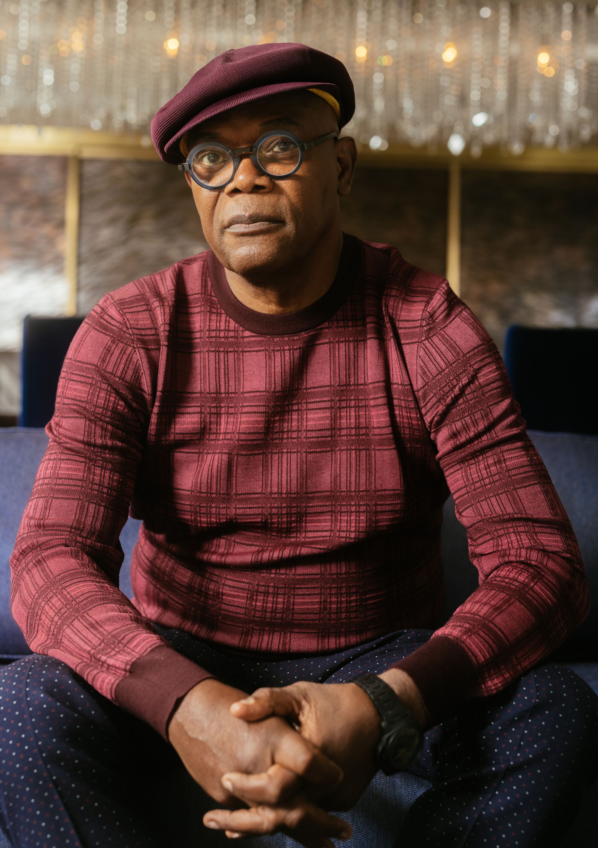 A man in a burgundy sweater sits on a couch with his hands clasped.