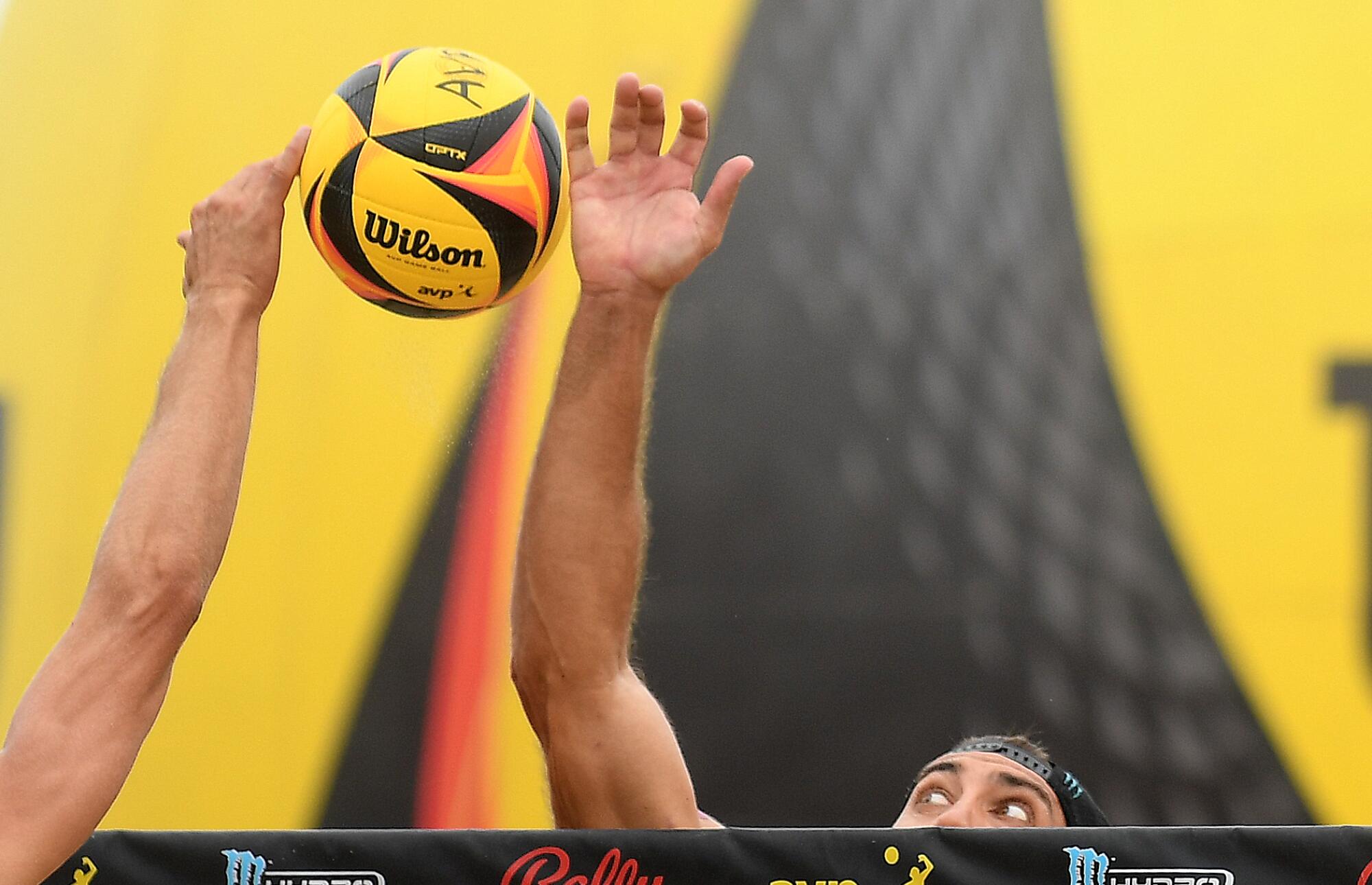Volleyball players' hands reach over the net to try to hit a yellow and black Wilson volleyball.
