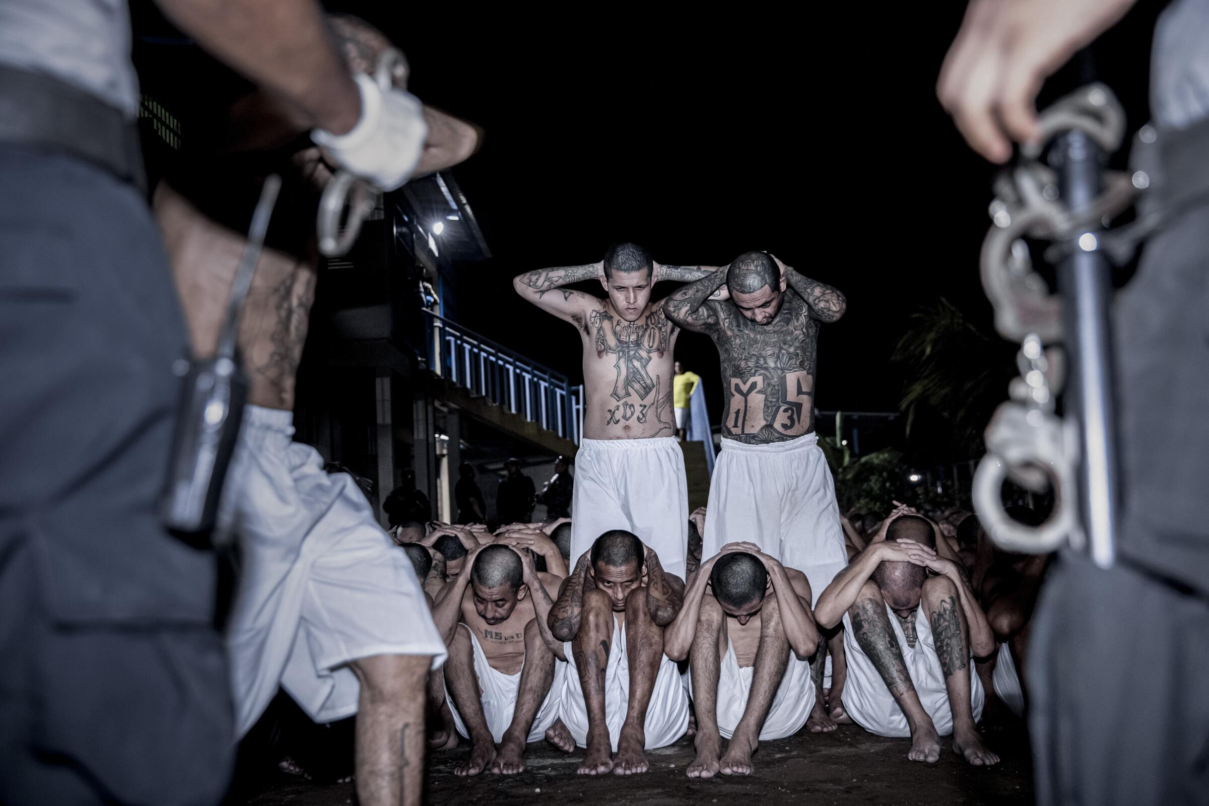 3 tattooed men in white boxers on their feet at night, hands behind their heads, as more sit facing 2 officers, foreground