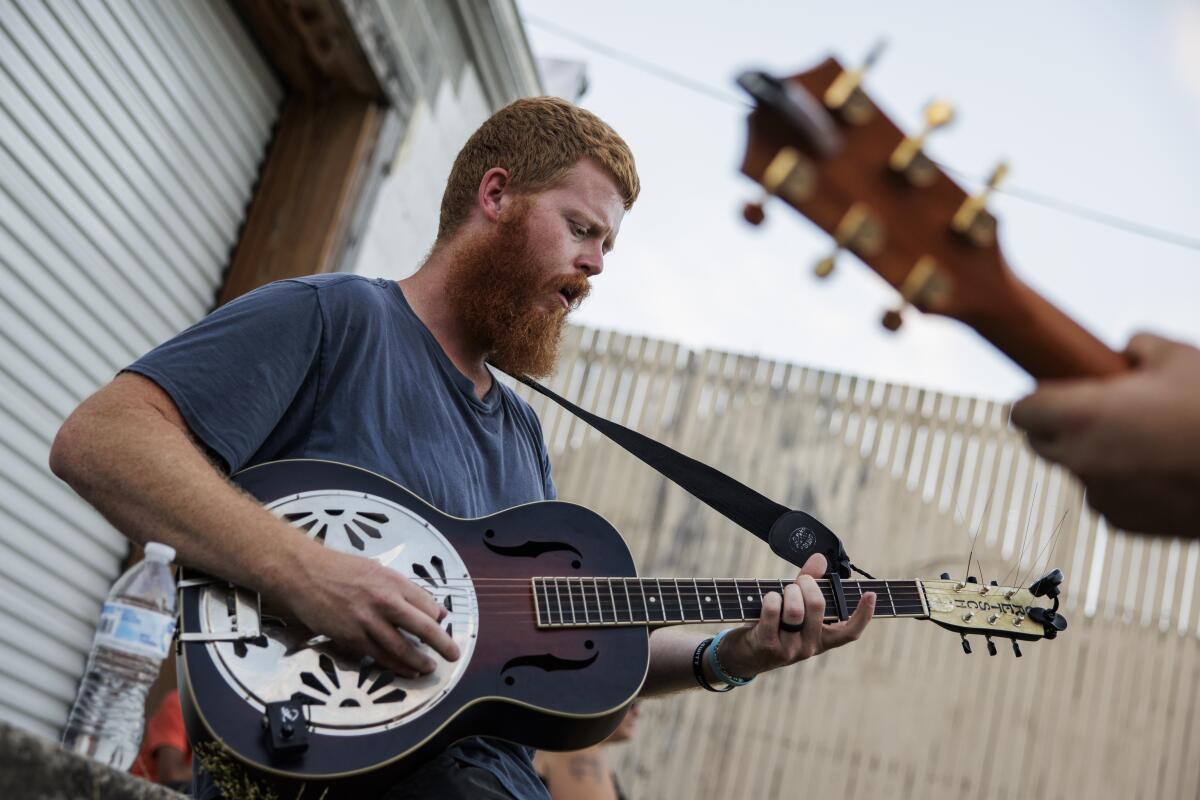 Oliver Anthony, with red hair and a full beard, plays a guitar. 