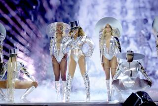 Beyoncé in a silver body suit, hat and boots singing on a stage surrounded by backup dancers in similar attire