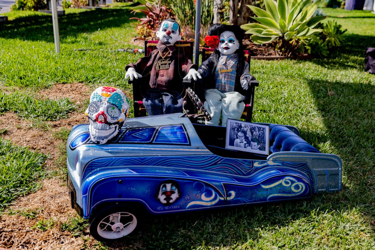 Two dolls with clown face paint sit in front of a mini vintage blue car displaying a sugar skull and a picture frame.