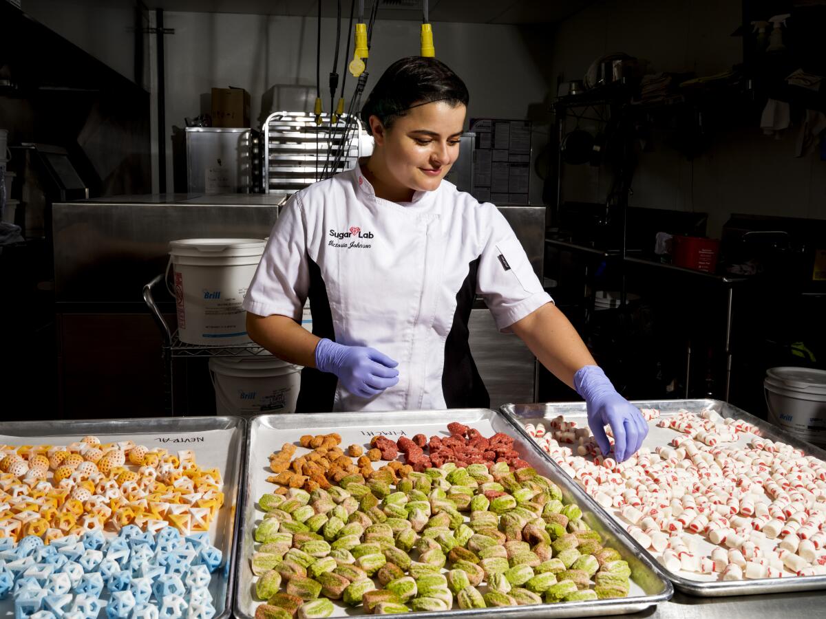 A woman in a chef's outfit and rubber gloves sorts through colorful trays of sugar candies and savory bouillon.