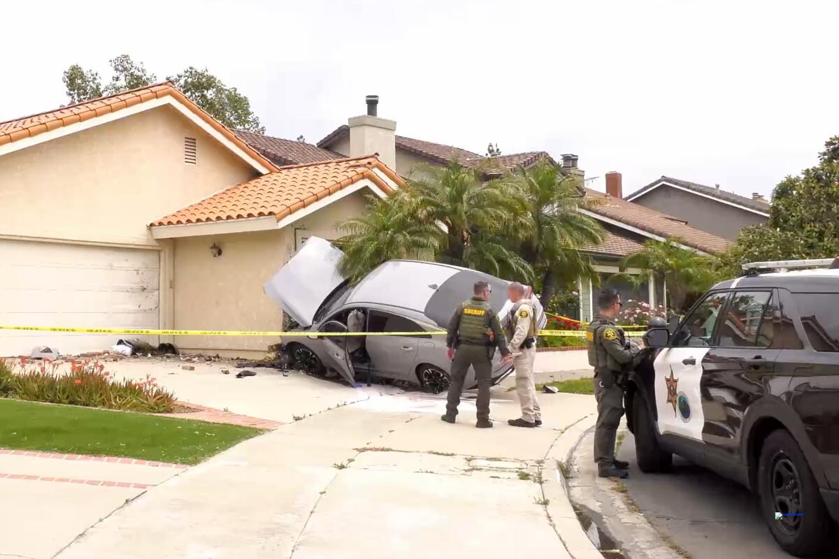 Three men in sheriff's uniforms standing outside a house surrounded by yellow police tape, with a car crashed into its side.