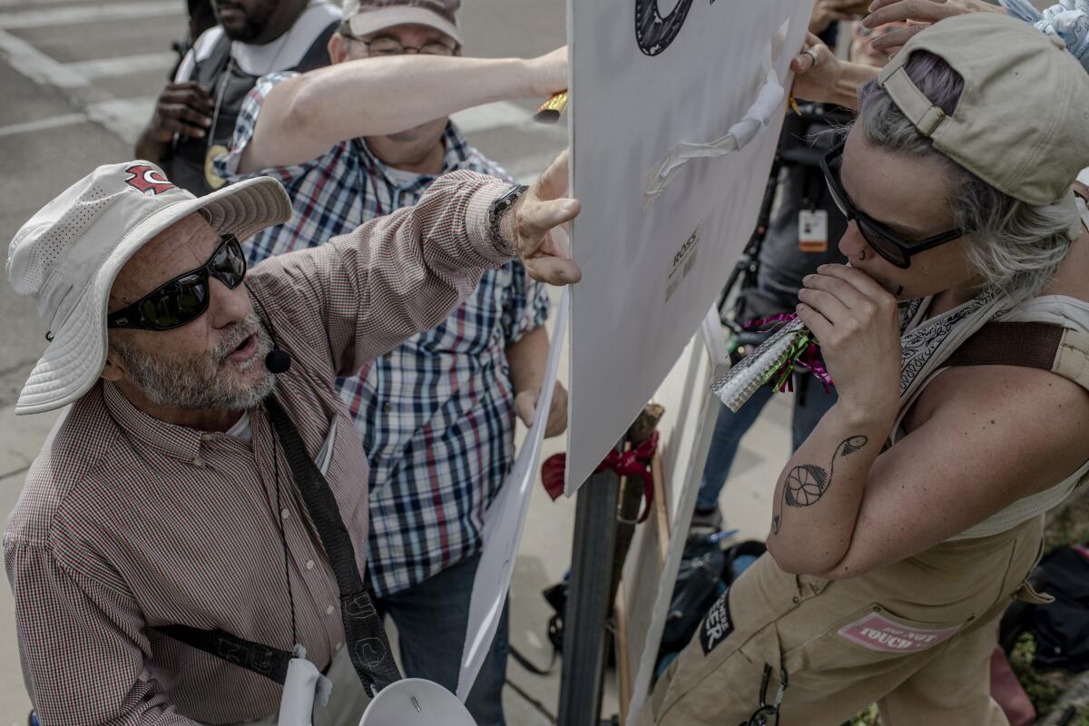 A man in a hat, separated by a sign, faces a woman holding a noisemaker 