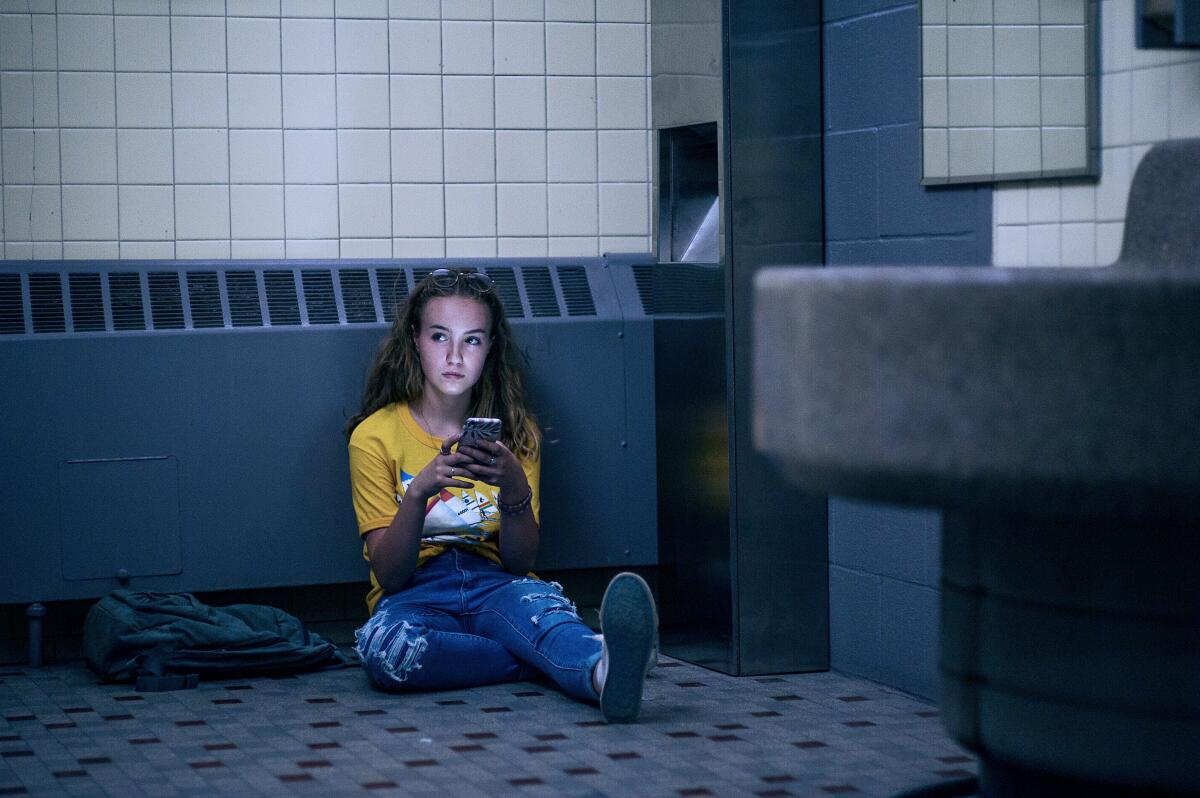A teenage girl sits on the floor of a restroom texting on her phone.