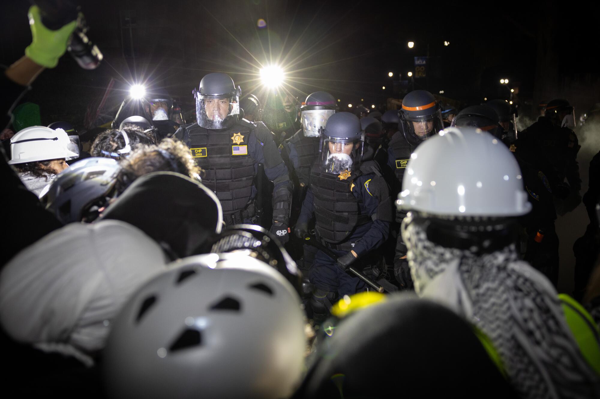 Police face people in white hard hats at night.