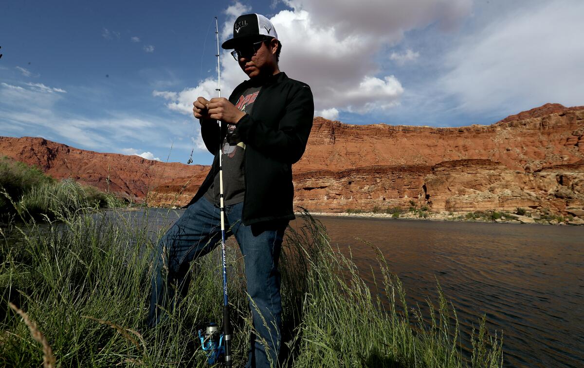 A person stands amid grass next to water backed by red cliffs; holding rod and reel, he baits a hook.