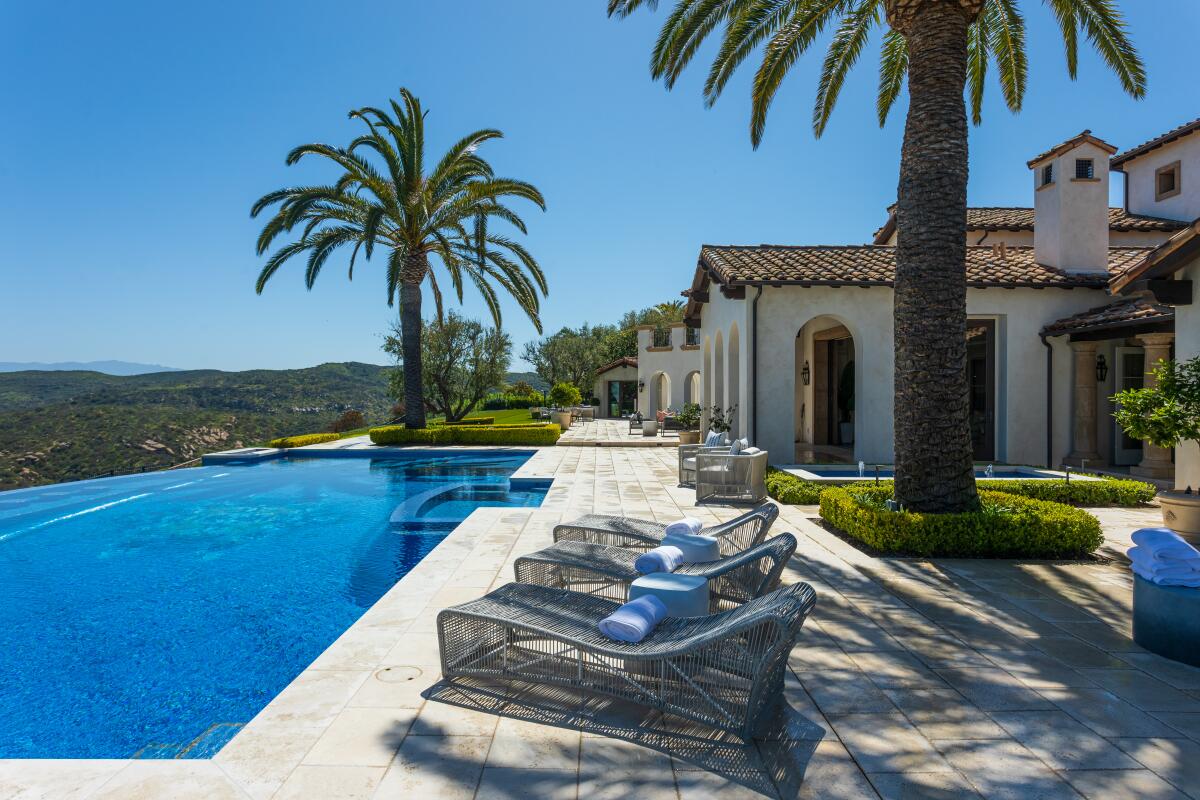 Lounge chairs are lined up next to a pool outside a Mediterranean-style mansion