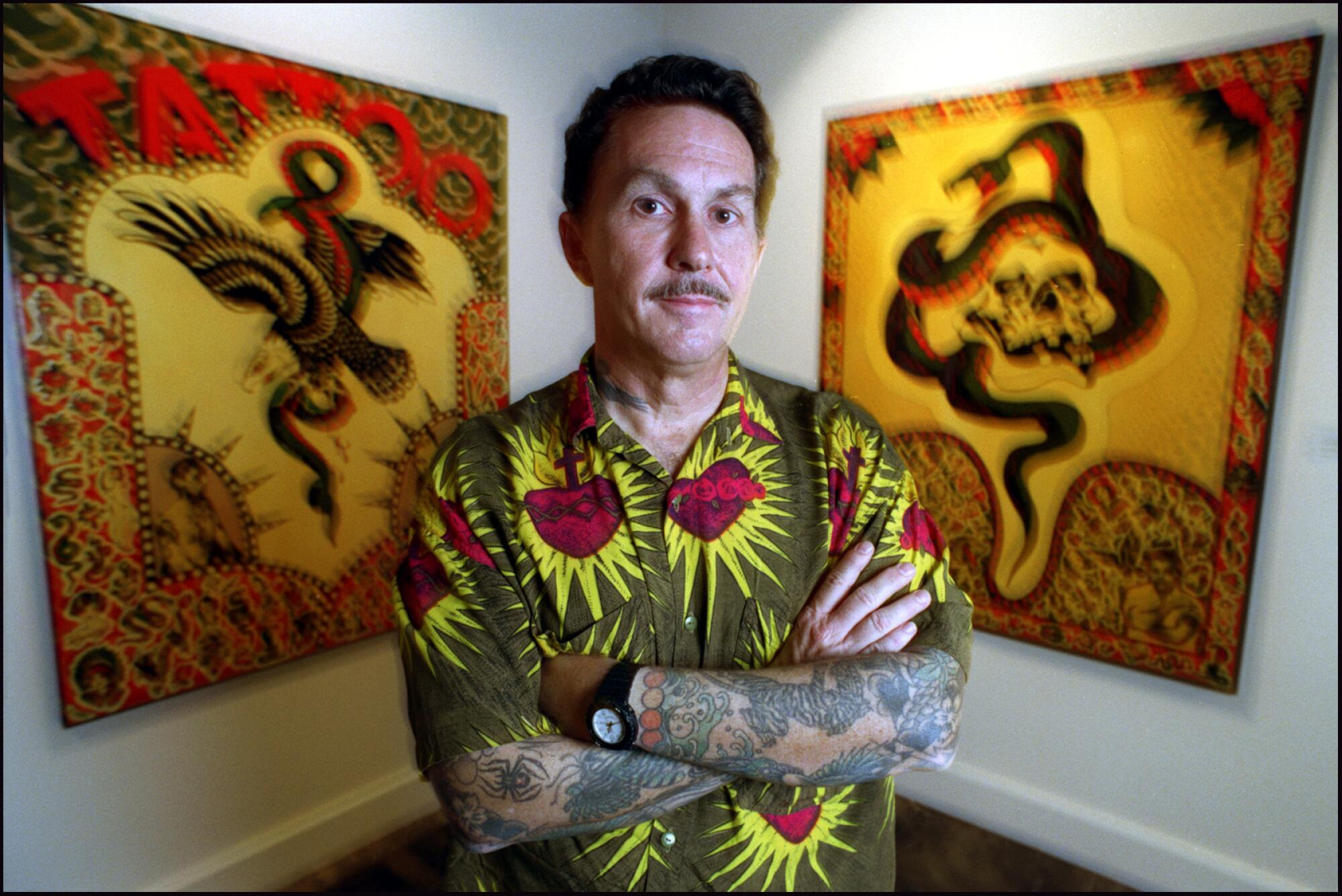 Don Ed Hardy stands before two panels of tattoo art, which also covers his shirt and both arms.