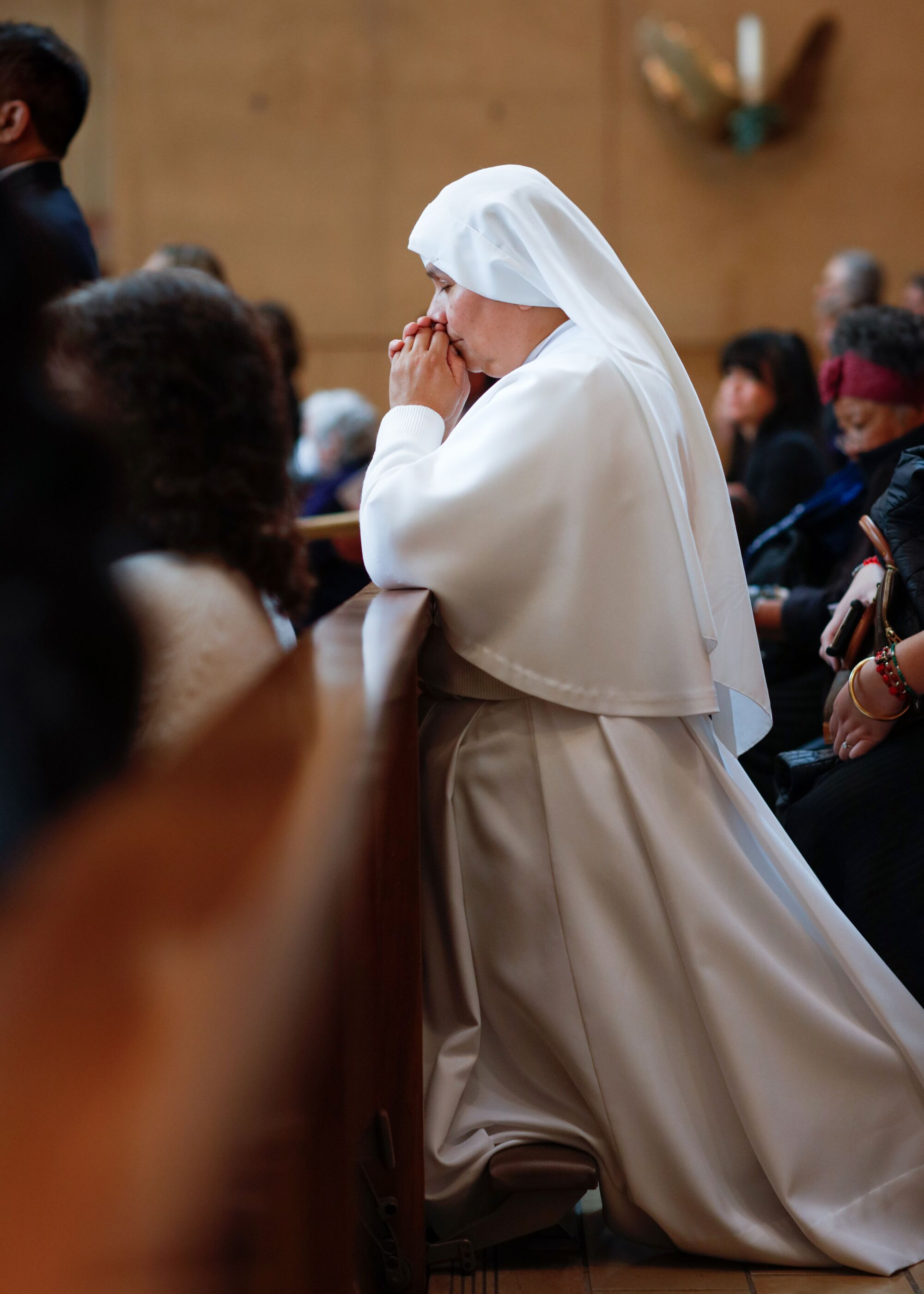 A nun in white habit kneels and prays in church pews