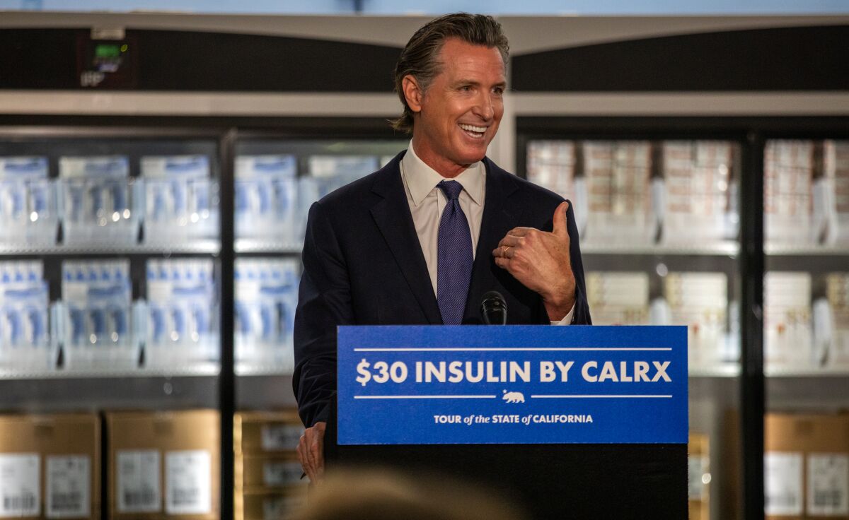 Gavin Newsom gesturing at a lectern near glass-doored refrigerators filled with boxes of insulin