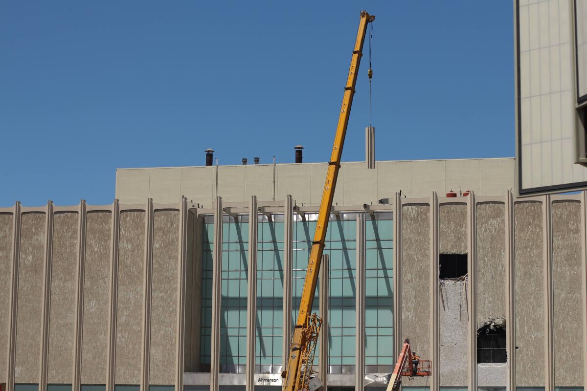 A bright yellow construction crane hoists a vertical piece of column from LACMA's facade into the air.