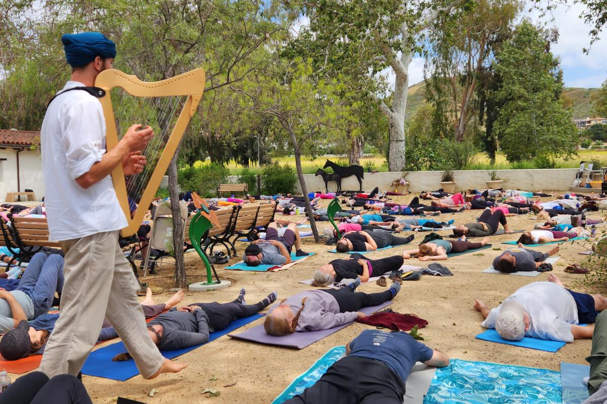 A standing person plays a small harp among dozens of people lying on yoga mats on the ground