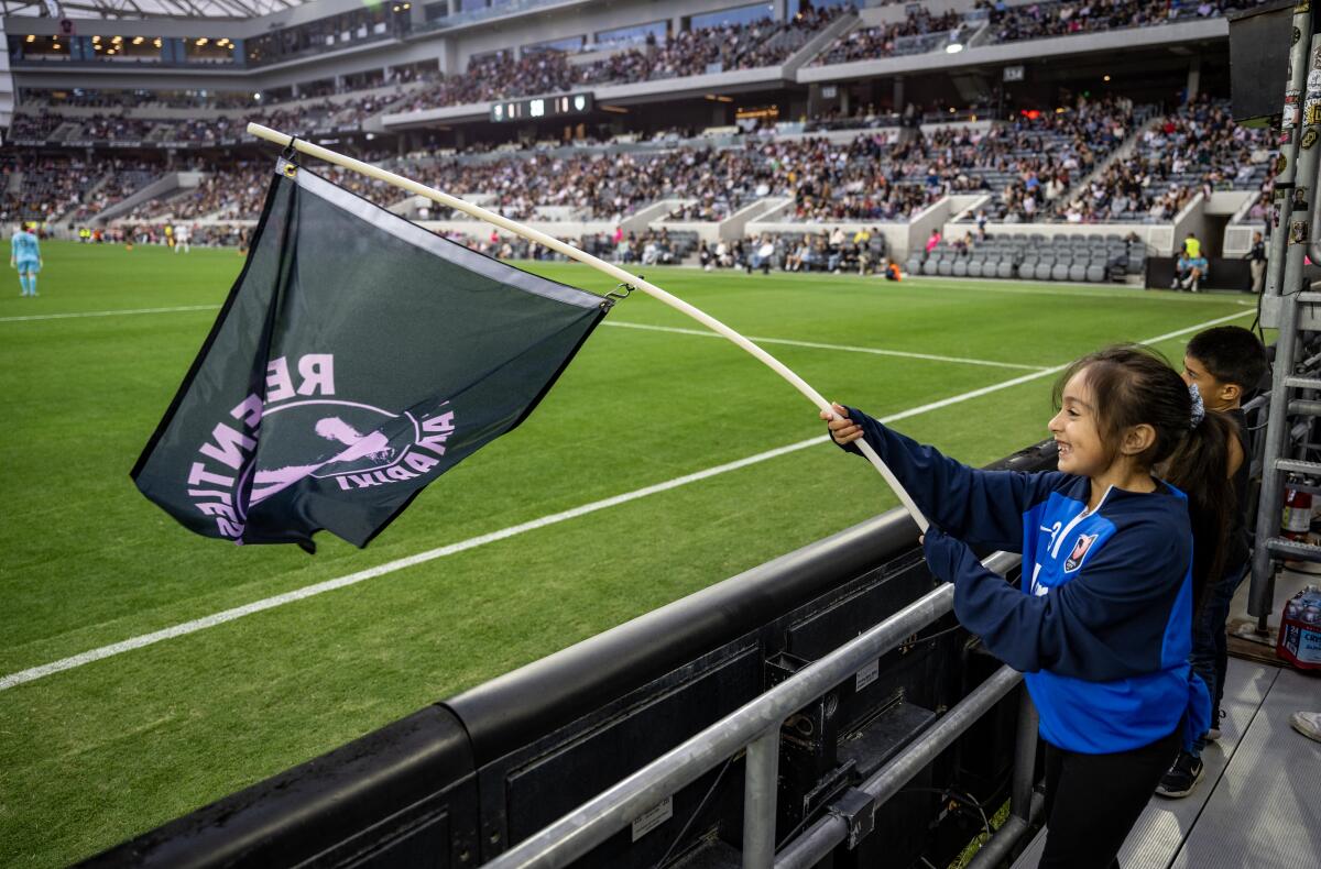 A young girl standing near the sidelines of a soccer field waves a large green flag.