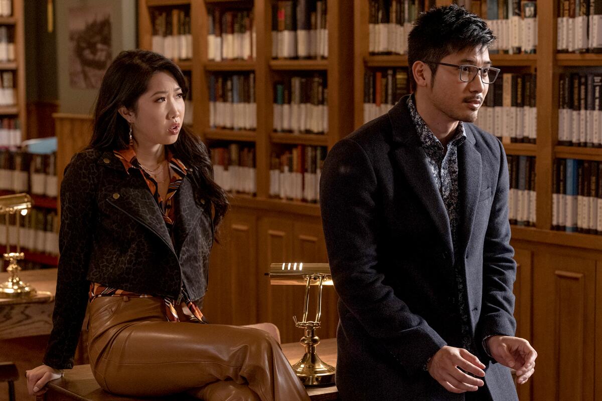 A man stands and a woman sits on a desk in a library.