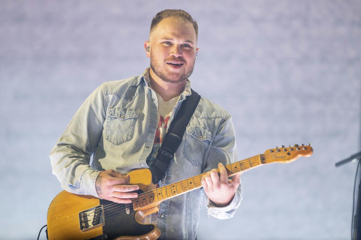 Zach Bryan in a T-shirt and denim button-down strumming a guitar during a performance on a stage