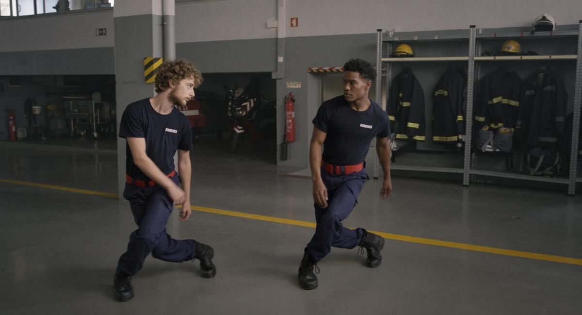 Two men strike a dance pose in a firehouse, with uniforms hanging in cupboards behind them.