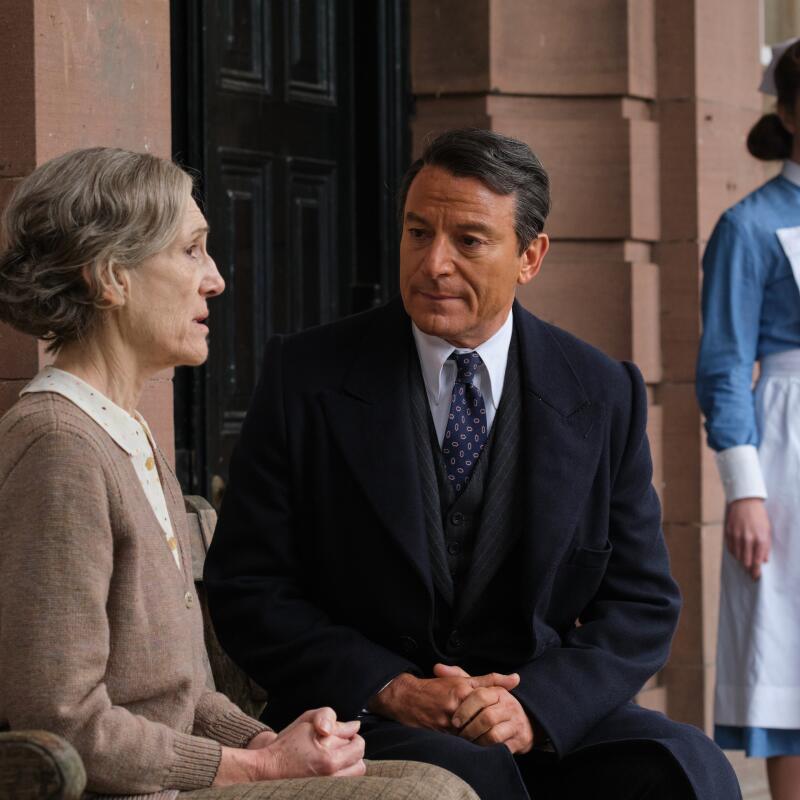 An older woman in a cardigan sits on a bench next to a man in a dark suit. A uniformed nurse stands nearby looking at them.