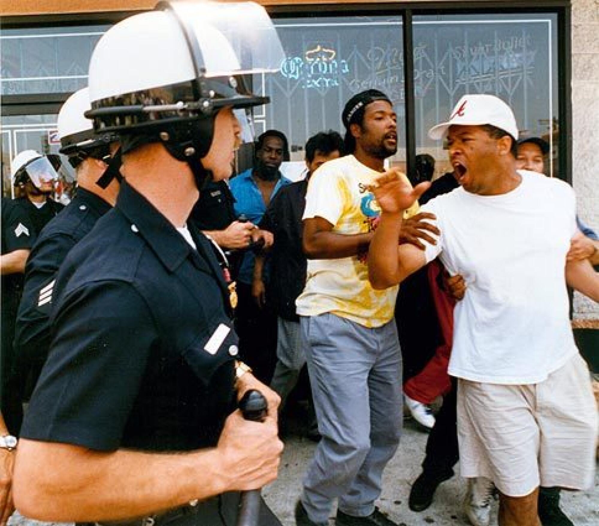 Two people hold the arms of a man who yells in the direction of a line of helmet-wearing police.
