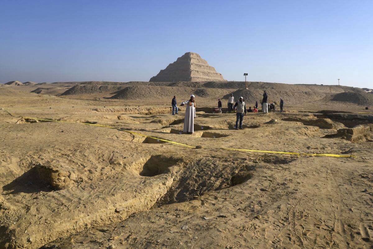 People stand in a desert landscape, with yellow tape and holes in the ground, and a pyramid as a backdrop 