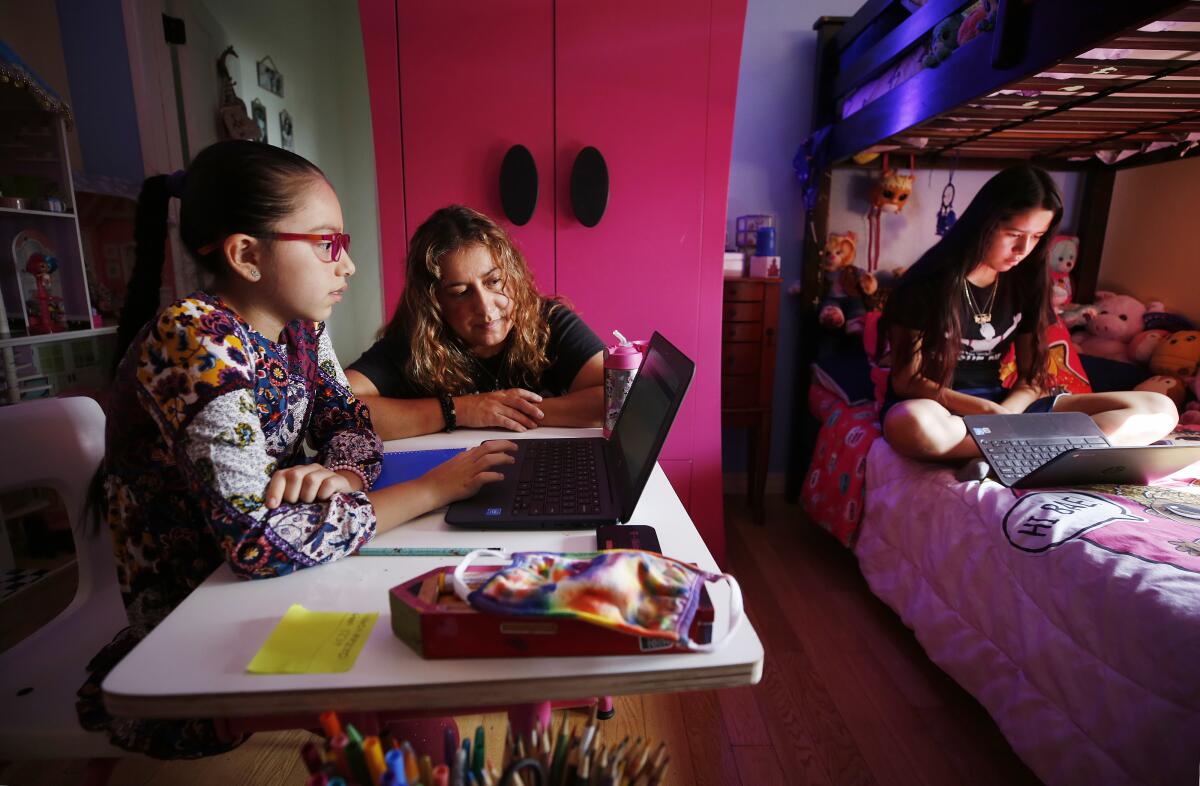 A mom helps her daughter working on a laptop at her desk, as her other daughter works on the bed