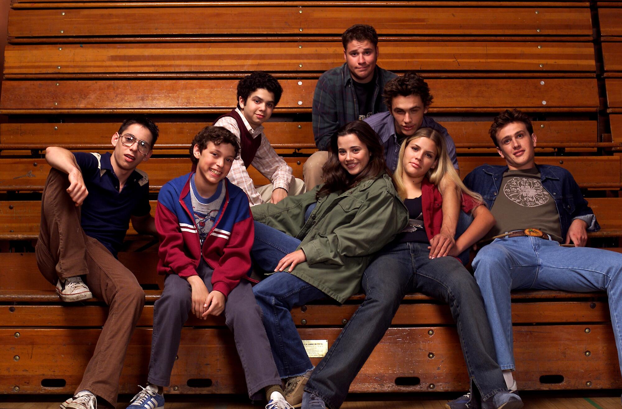 Eight teenagers sitting on wooden bleachers in a gymnasium.