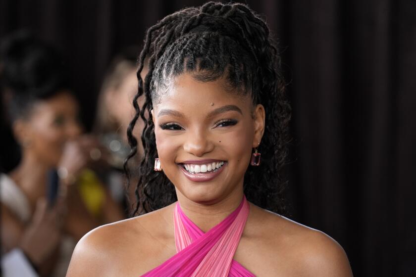 Halle Bailey in a pink halter dress with her hair in a high ponytail smiling an posing against a black background