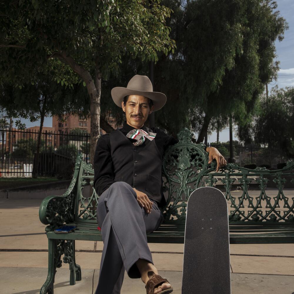 A man in ranchero-style clothing sits on a bench with a  skateboard at his side
