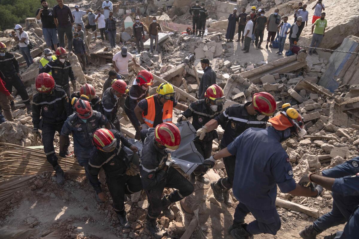 People in red hard hats and dark uniforms carry a body, with other people gathered amid rubble 
