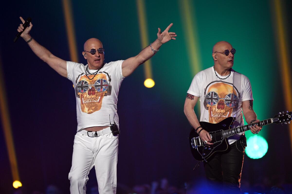 Two male musicians with matching bald heads, sunglasses and skull T-shirts perform onstage, one playing a guitar.