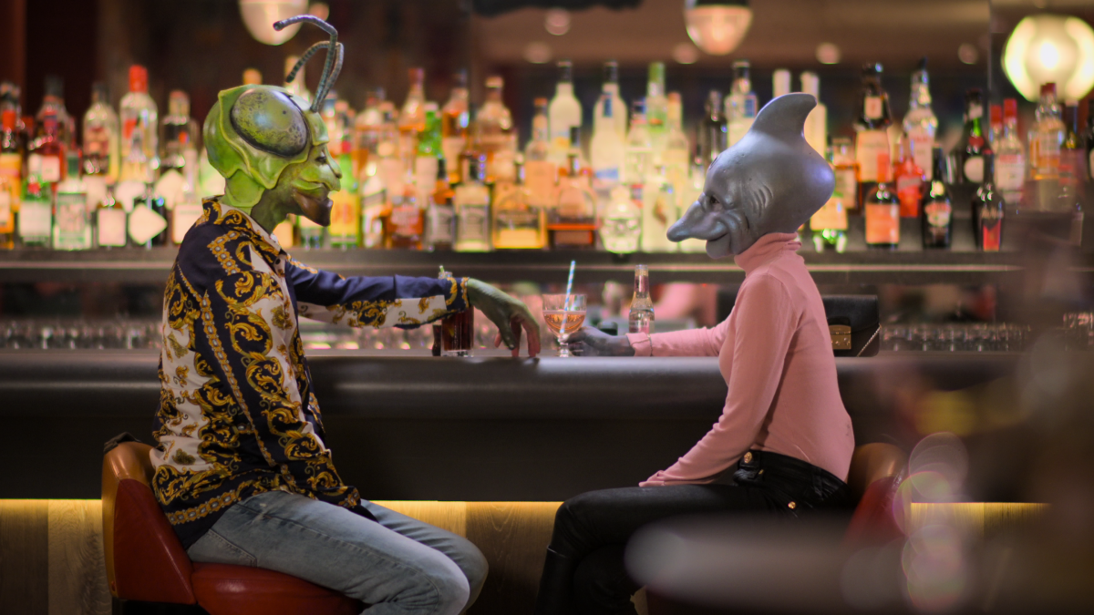 A grasshopper-headed person and a shark-headed person dressed in regular clothes have a drink at a bar.