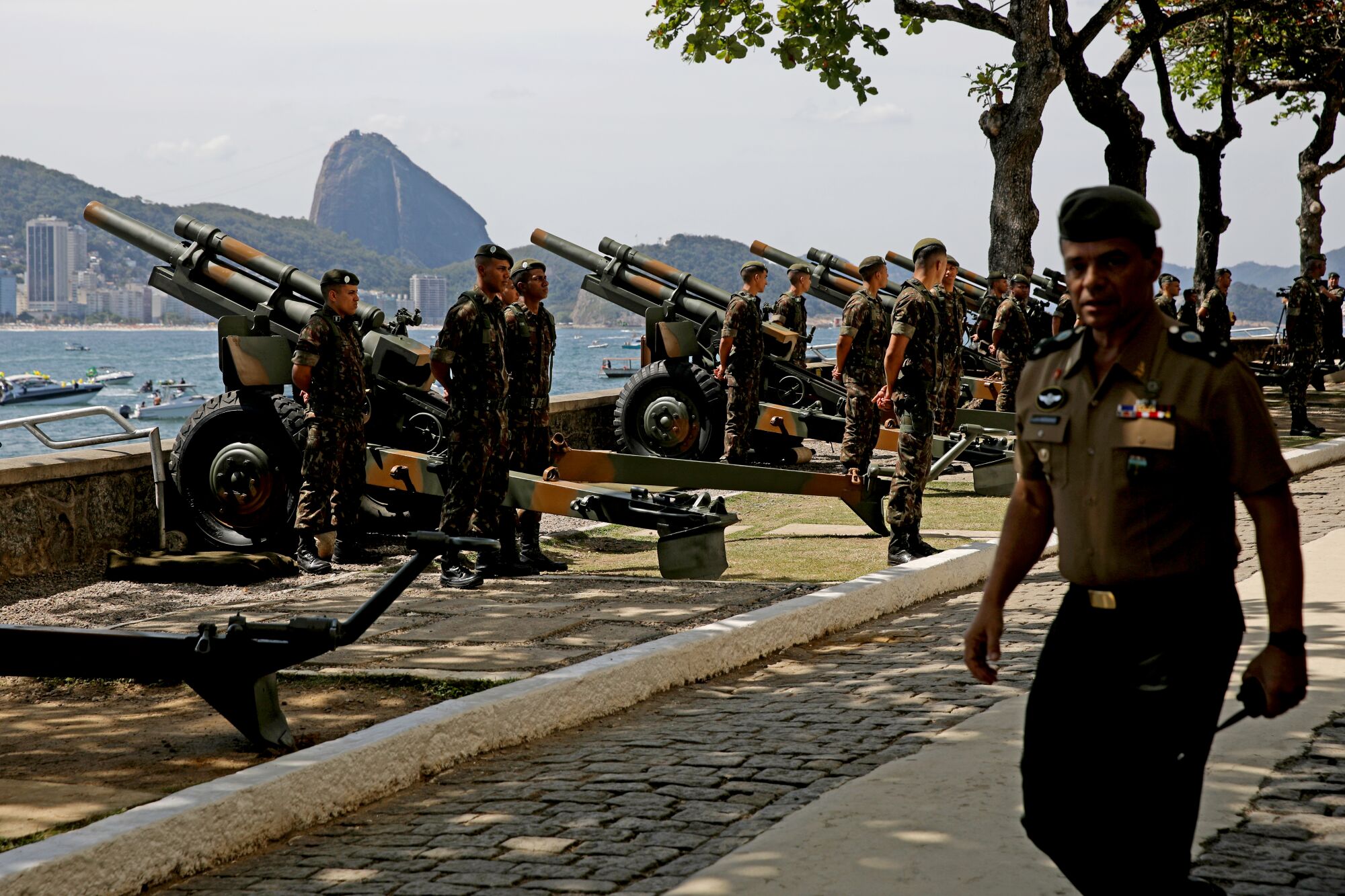 A man in a beret and military uniform passes other people in fatigues standing near cannons with the ocean as a backdrop