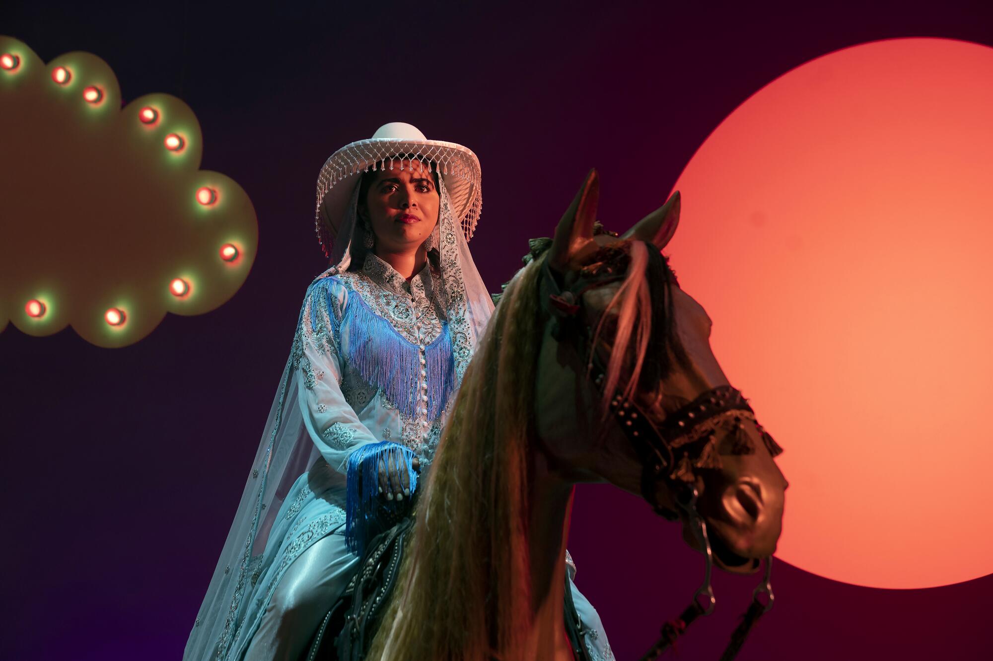Malala Yousafzai sitting on top of a brown horse wearing a cowboy shirt and hat with beads hanging down the brim.
