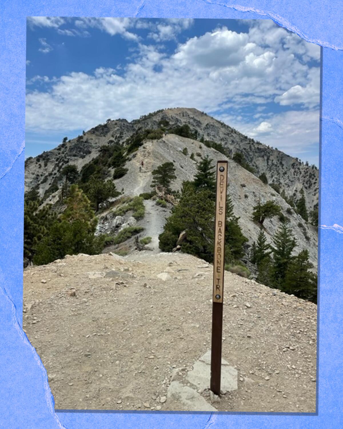 A narrow marker stands on a peak. In the background is another mountain with low green trees, and blue sky and clouds.