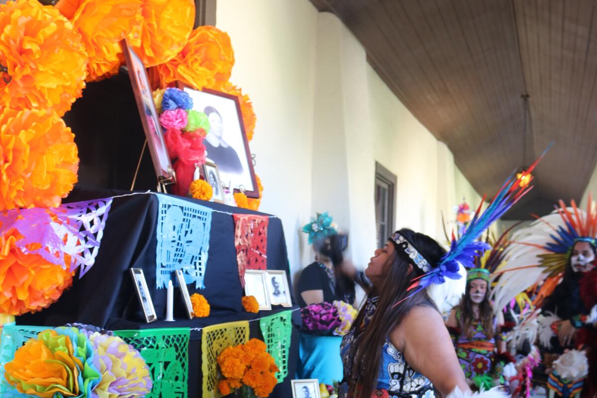 A woman in traditional clothing stands in front of an altar decorated with papel picado. 