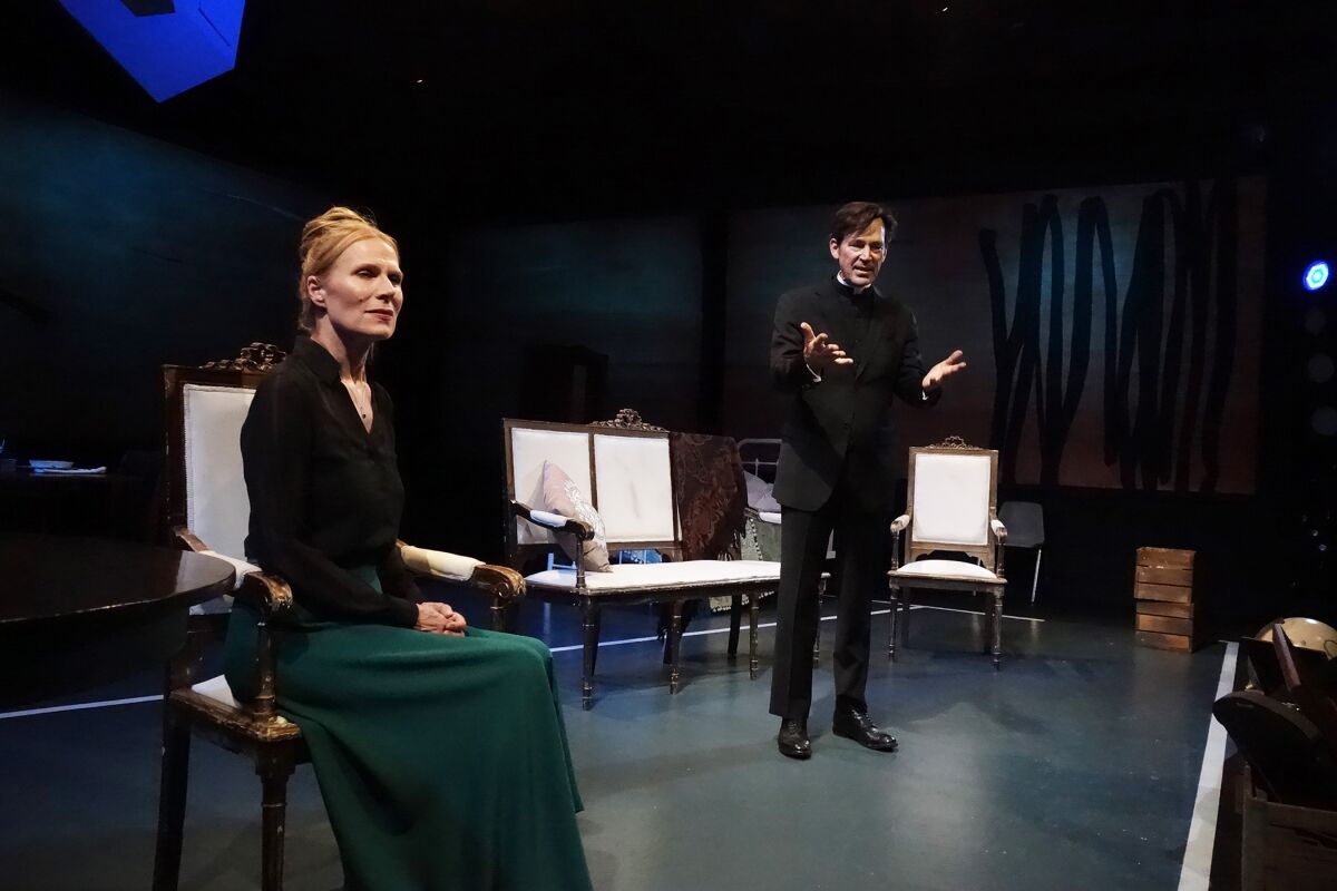 On a stage, a man points to a seated woman.