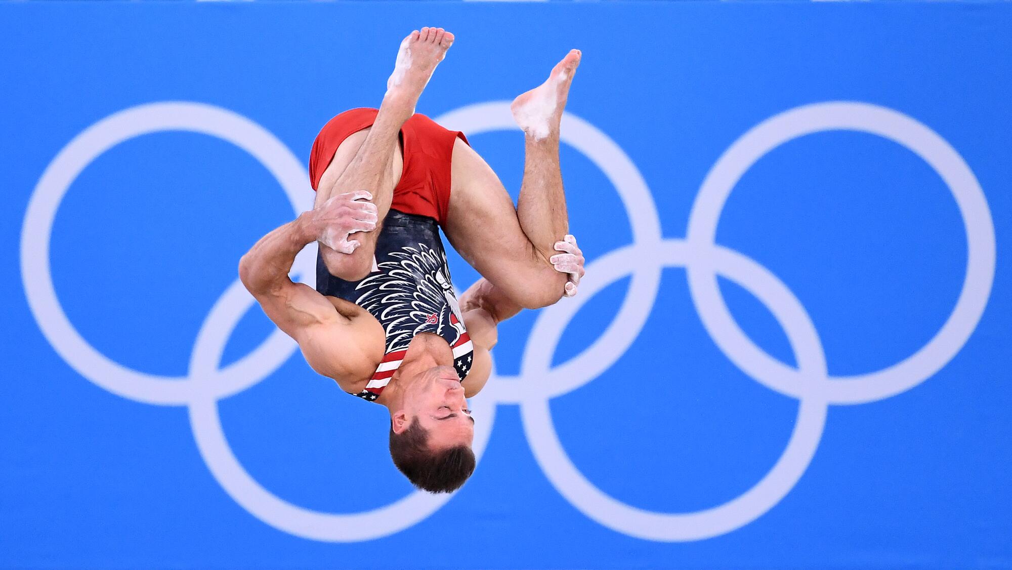 Samuel Mikulak is upside down, grasping both knees, with the Olympic rings in the background.